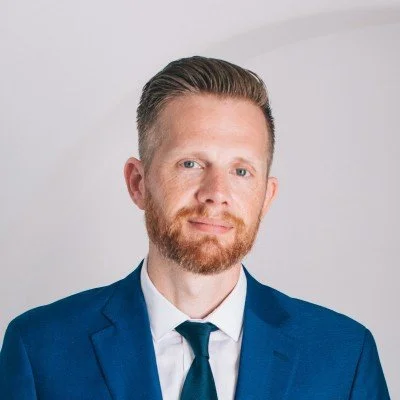 Professional portrait of a man with short, slicked-back hair and a beard, wearing a blue suit, white shirt, and dark tie, with a plain white background.