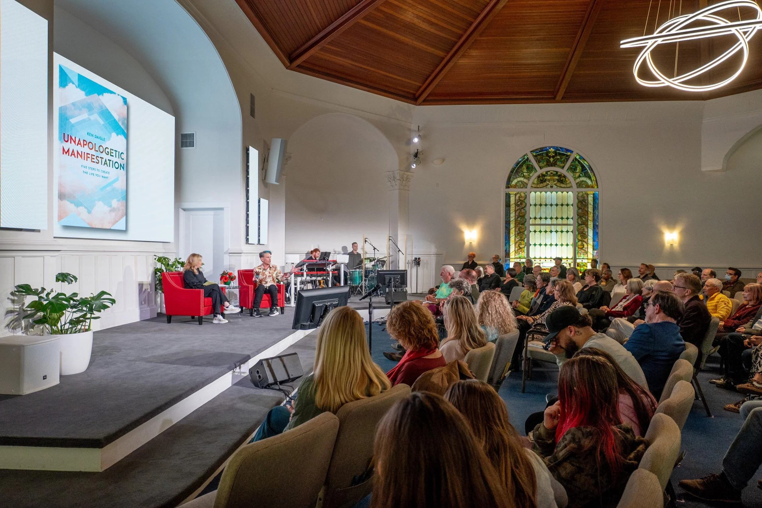 An indoor event with a stage and a seated audience. The stage has a woman speaking, along with a man and a small musical setup. The audience sits in chairs facing the stage, with some people taking notes and others attentively listening. The venue has high ceilings with wooden panels, stained glass windows, and modern lighting fixtures.