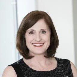 A woman with shoulder-length brown hair smiling, wearing a black sleeveless top with a subtle pattern, standing in front of a light-colored background.