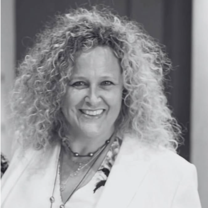 A smiling woman with curly hair wearing a blazer, necklace, and a patterned blouse.