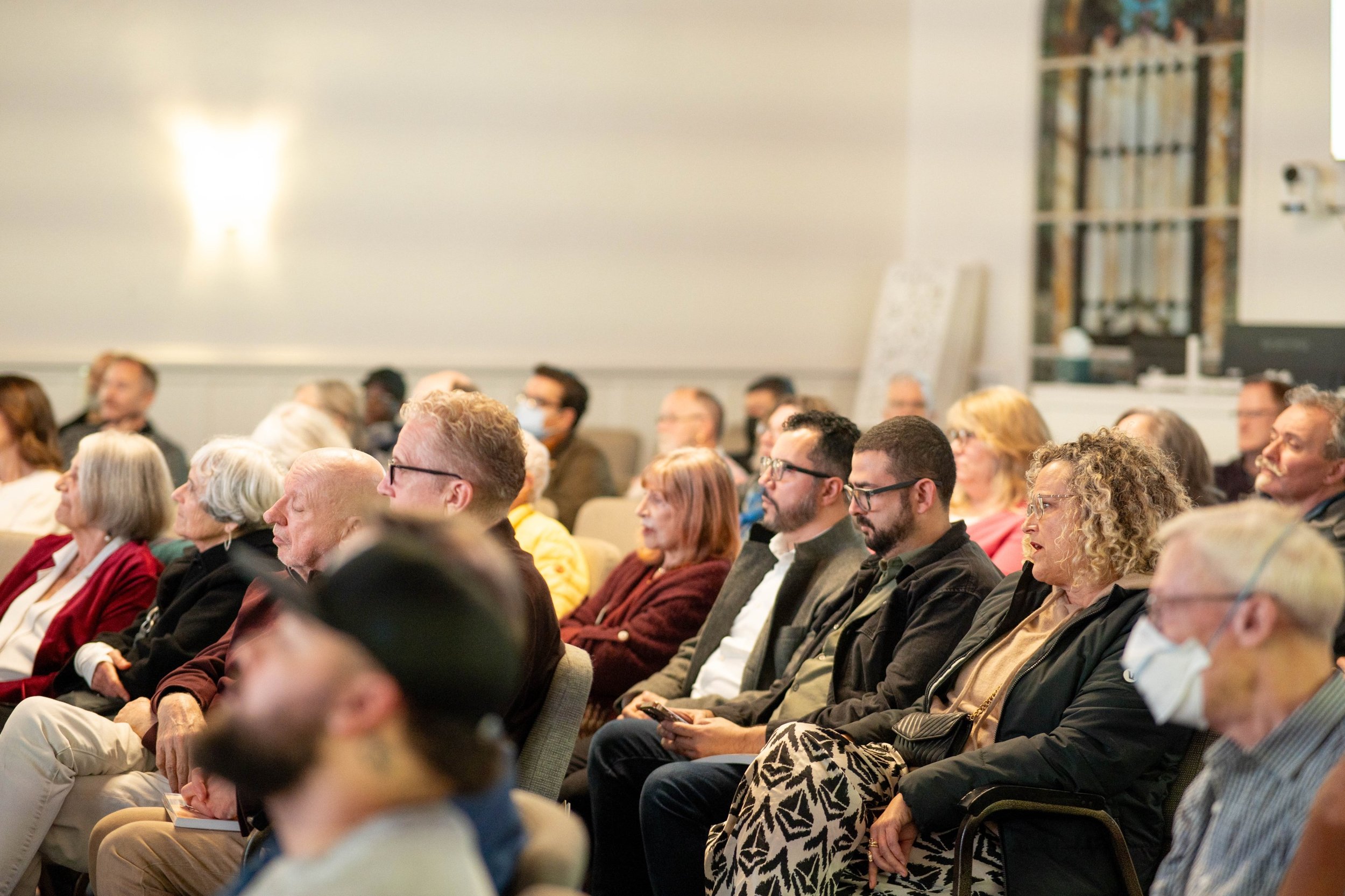 A diverse group of adults seated closely together in an indoor conference room or auditorium, attentively listening to a presentation.