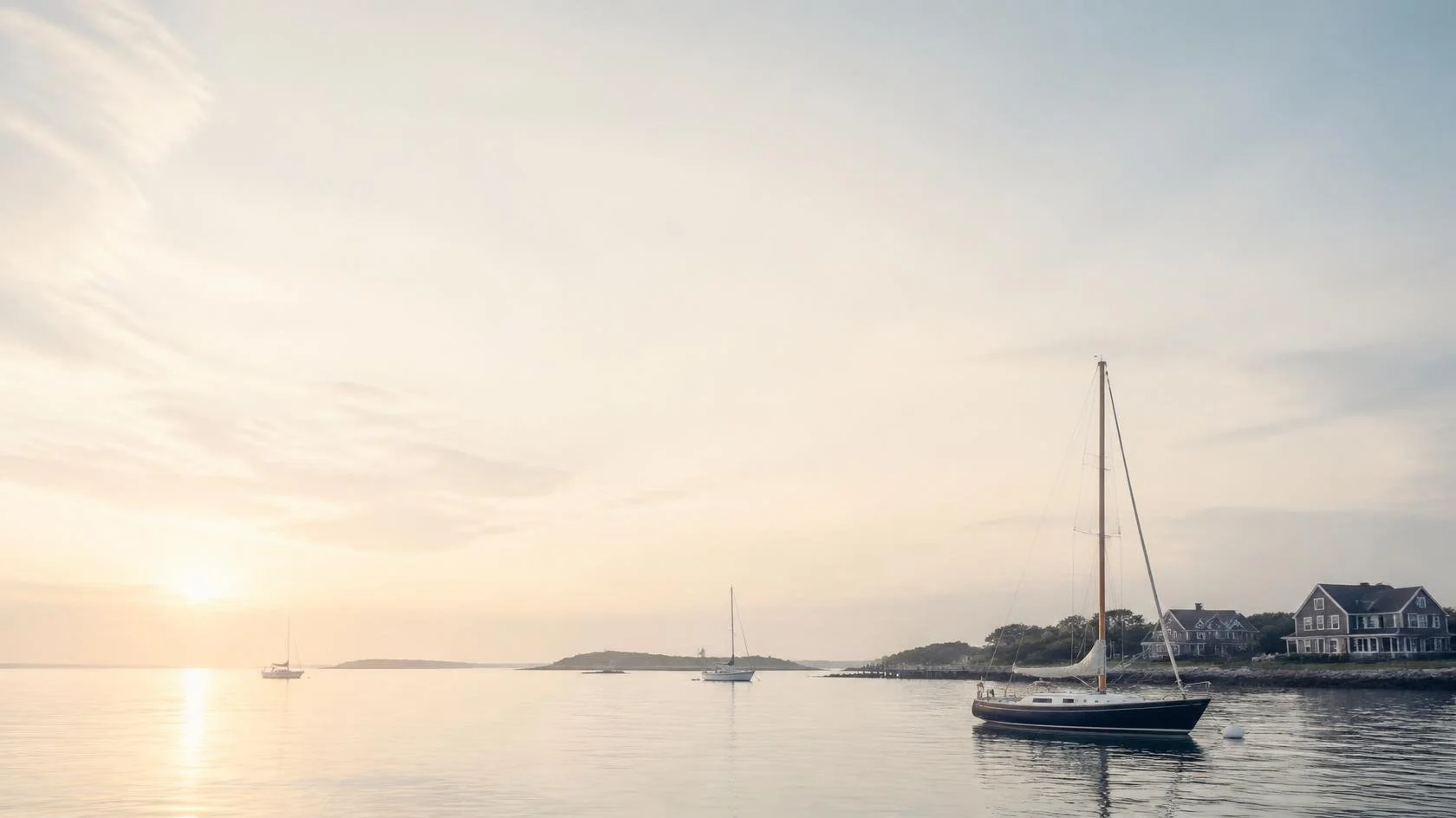 A serene waterfront scene at sunset with sailboats on calm water, houses on the shoreline, and a partly cloudy sky.