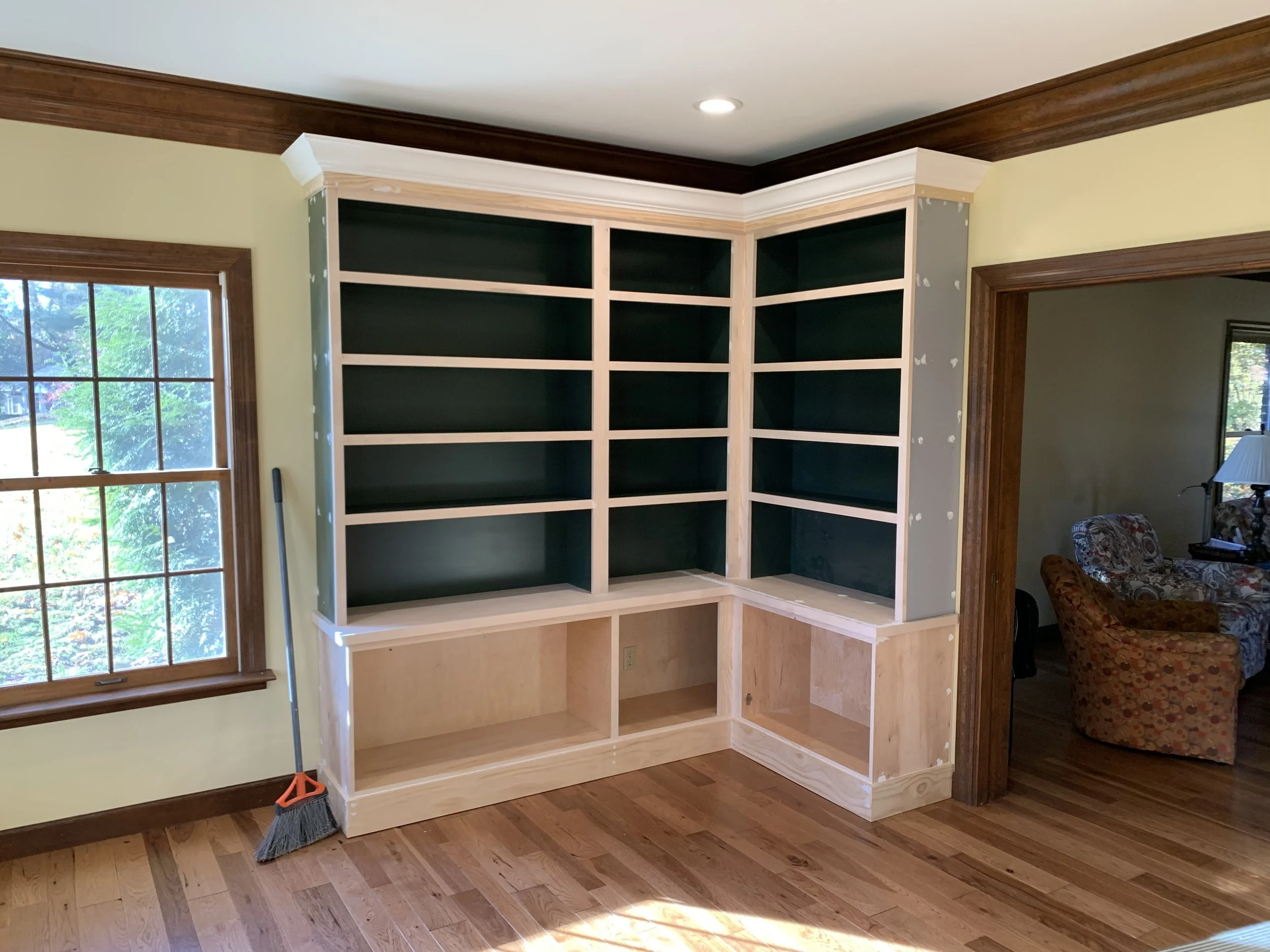 A partially built wooden bookshelf unit against a wall in a room with hardwood floors, a window on the left, and an open doorway on the right.