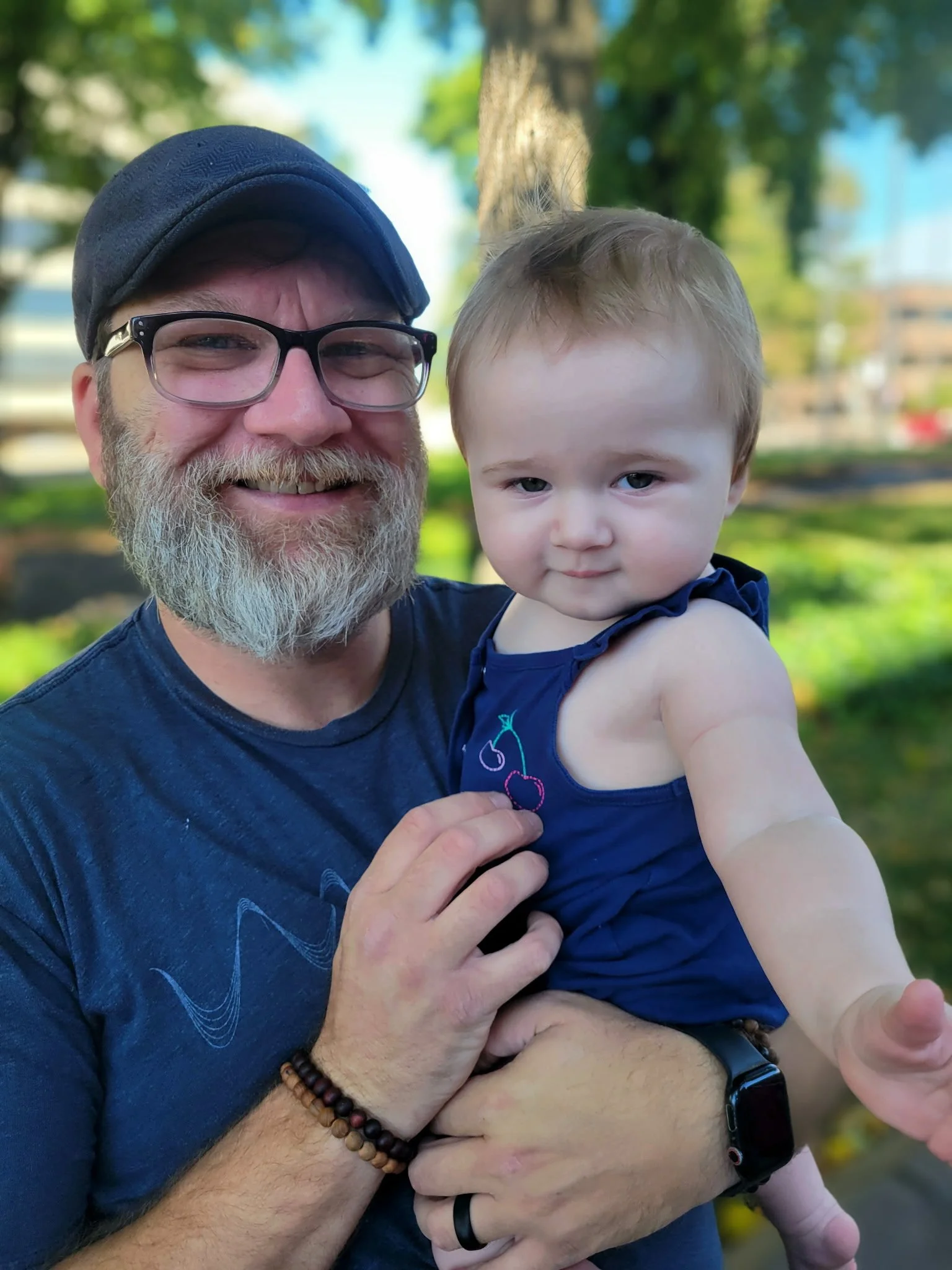 A smiling man with glasses and a beard holding a baby girl outside in a park with trees and buildings in the background.