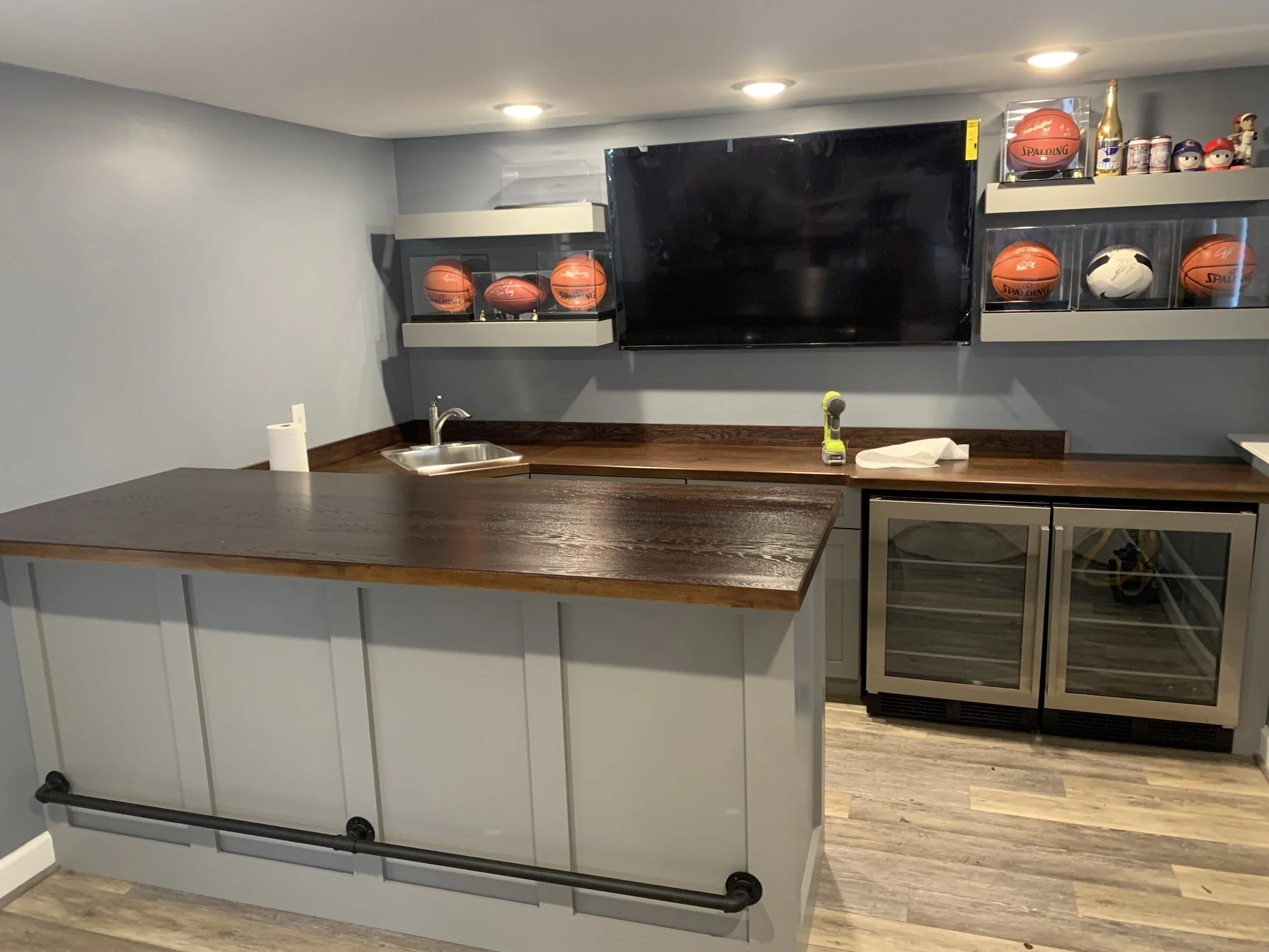 A basement kitchen area with a wooden countertop, a stainless steel sink, a paper towel roll, a small cordless drill, and a paper towel on the counter. The wall has two shelves housing basketballs and other small collectibles, and a large flat-screen TV is mounted on the wall. There is a mini refrigerator and an oven below the counter. Recessed ceiling lights illuminate the room, and the flooring is wood.