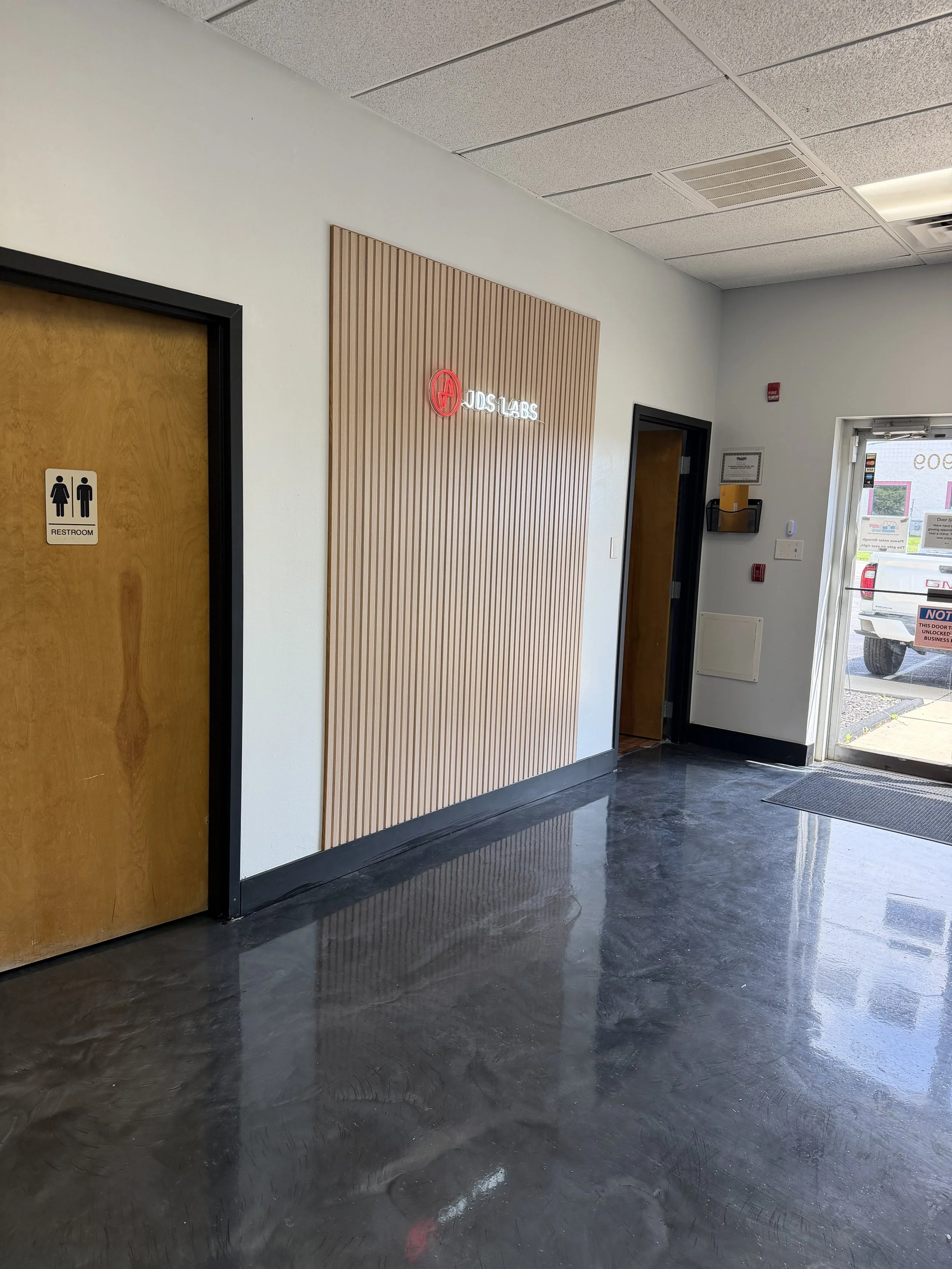 Interior of a commercial building, showing a wooden door with a restroom sign, a textured wall with a neon sign that reads 'OIDS LABS,' and a glass door at the entrance with a vehicle outside.