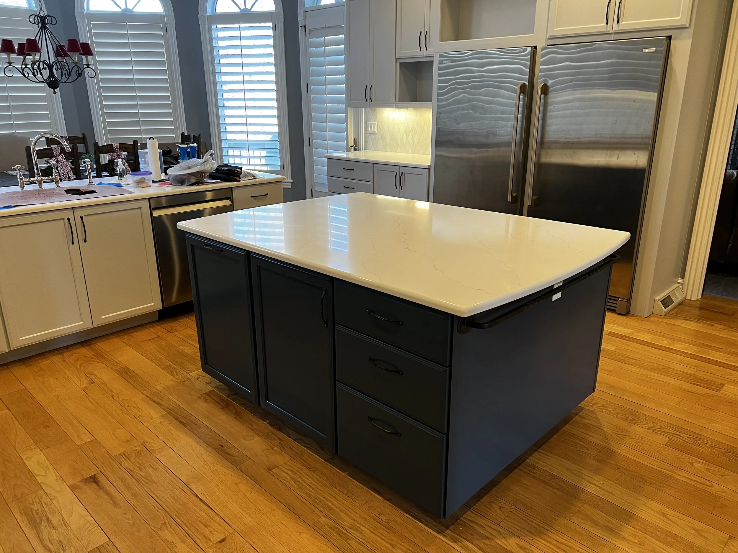 A modern kitchen featuring a large island with a white countertop and dark blue cabinetry, stainless steel appliances including a double-door refrigerator, white cabinetry along the walls, and large windows with white shutters, hardwood flooring, and a black chandelier with red lampshades hanging from the ceiling.