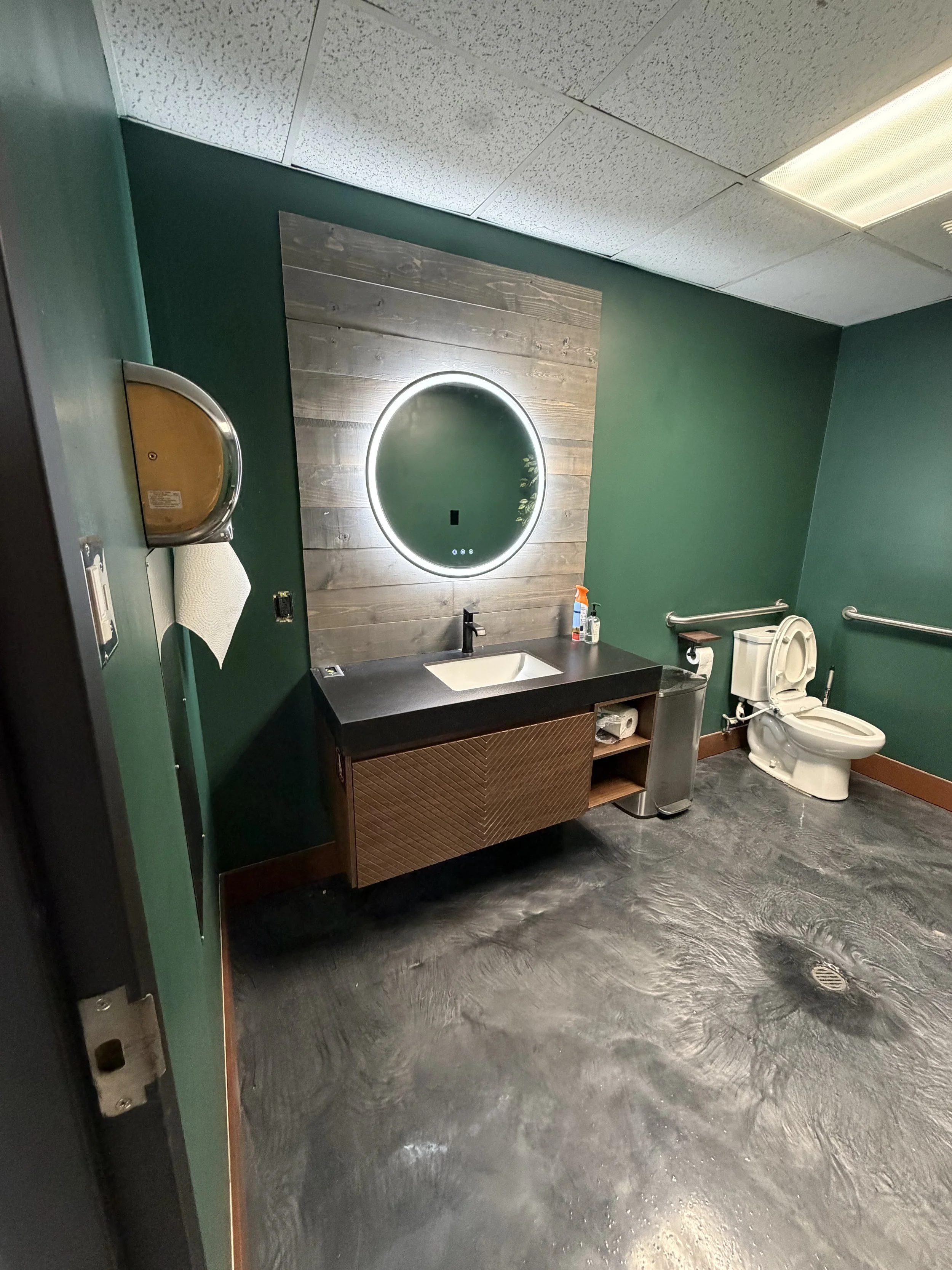 A bathroom with green walls, a wooden mirror with LED lighting, a black countertop with a sink, a soap bottle, a paper towel dispenser, a toilet with a toilet paper roll, a small trash can, and a floor drain.