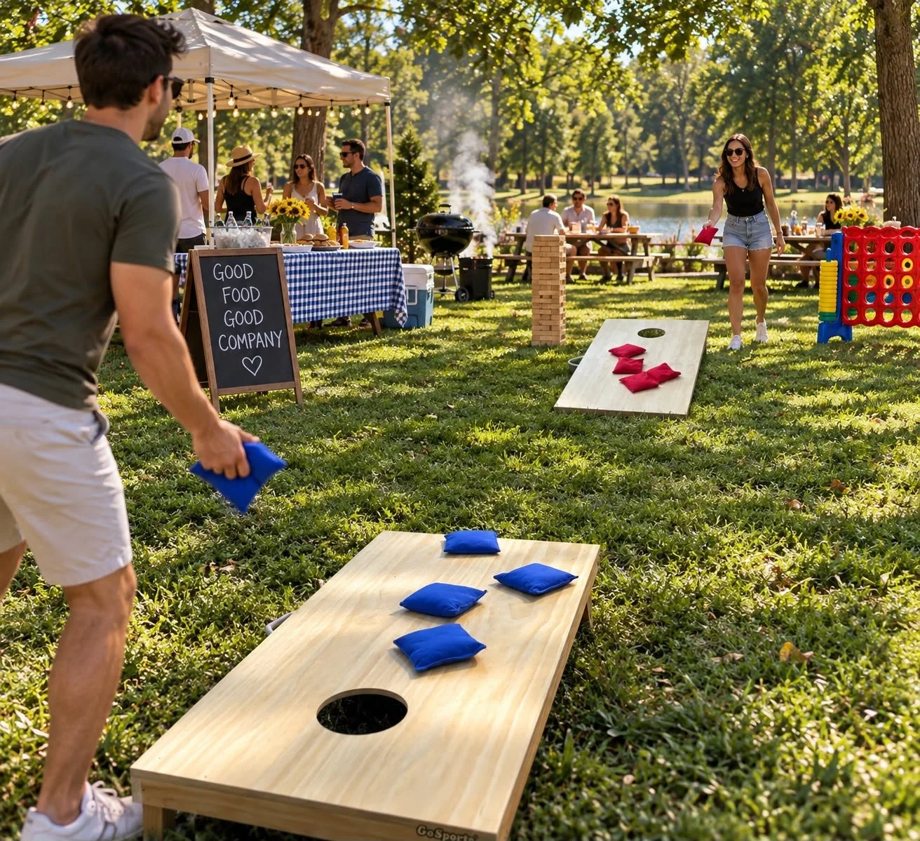 People playing cornhole games at a backyard gathering during daytime with a decorated table, a chalkboard sign, and several individuals in casual summer attire enjoying a sunny outdoor event near a lake.