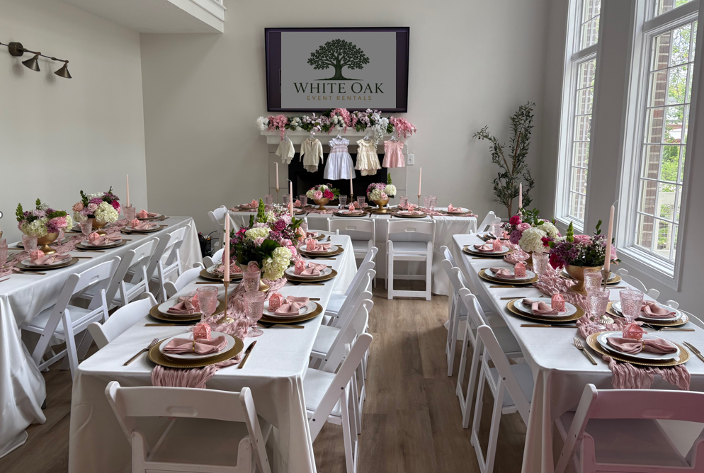 Elegant indoor event space decorated for a celebration with tables set with pink and white floral centerpieces, pink glassware, pink napkins, and gold-rimmed plates, with a large screen displaying 'White Oak Event Rentals' and a floral backdrop.