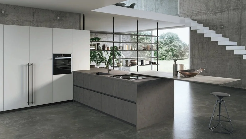Cucina moderna in stile minimalistico con pareti in cemento, mobili bianchi e isola centrale in cemento con piano in legno, scaffale aperto con libri e piante, grande finestra che mostra natura esterna, e una sedia da bar in metallo.