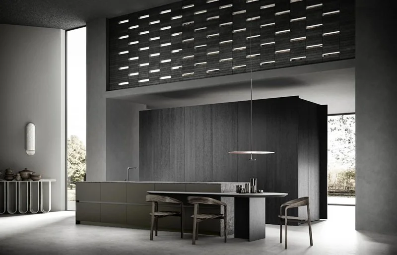 Interno di una cucina moderna con design minimalista, pareti in toni neutri, isola centrale con lavabo, tavolo con sedie in legno e grande finestra che lascia entrare molta luce naturale.