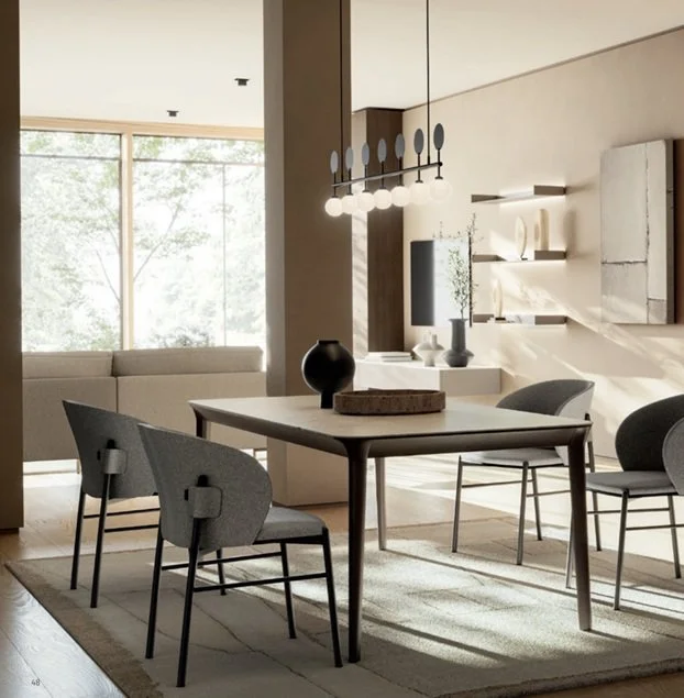 Interno di un soggiorno e sala da pranzo con mobili moderni, tavolo, sedie, lampadario e grandi finestre che lasciano entrare luce naturale.