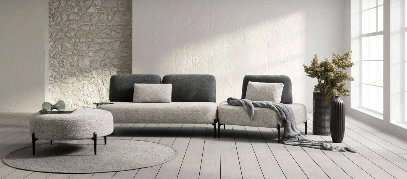 Soggiorno minimalista con divano grigio, pouf, tappeto, pianta in vaso e grande finestra con luce naturale.