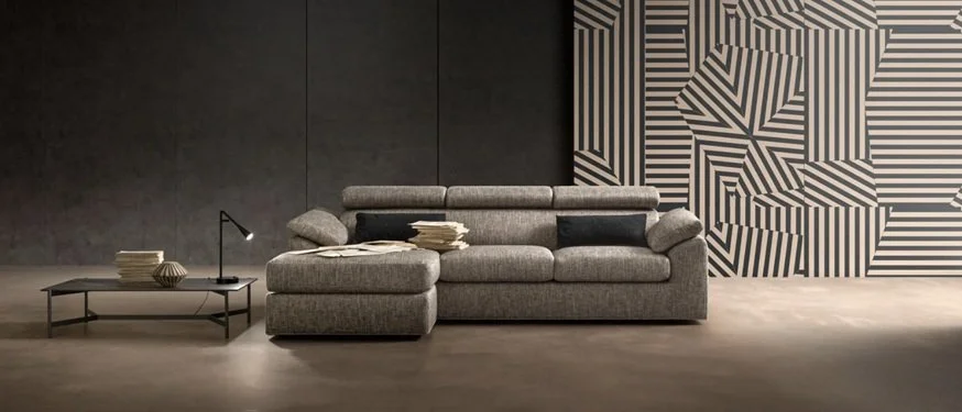Divano grigio in uno spazio moderno con un tavolino basso, una lampada da tavolo, vari libri e una parete decorativa con motivi geometrici