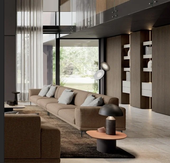 Interno moderno di un soggiorno con divano beige, grande finestra con vetri, mensole in legno con libri, lampada dal design contemporaneo, tappeto scuro, e arredamento minimalista.