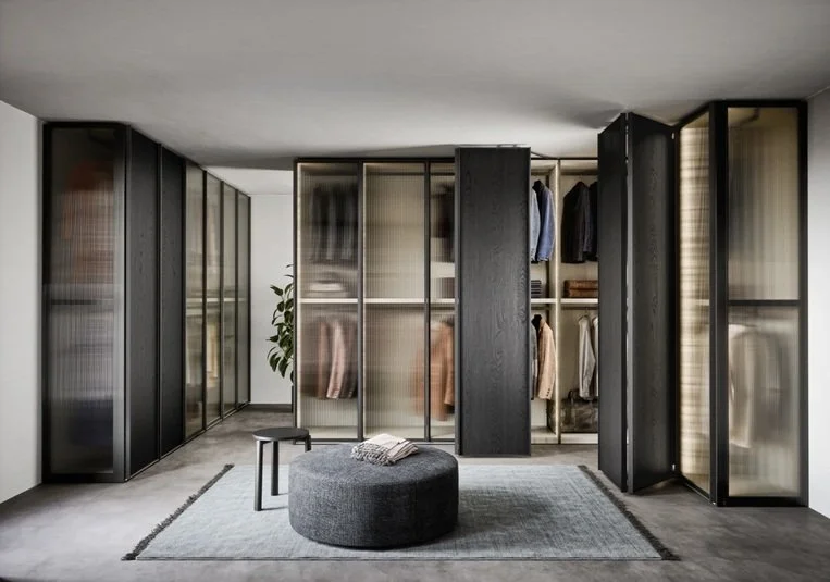 Armadio a specchio con ante scorrevoli e cabina armadio aperta con vestiti appesi, un tappeto grigio, un pouf rotondo e una sedia