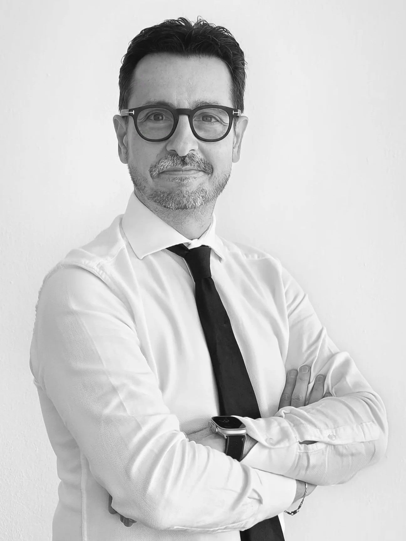 Ritratto di un uomo con occhiali scuri, capelli scuri e barba, che indossa una camicia bianca, una cravatta nera e un orologio smartwatch, che sorride leggermente e ha le braccia incrociate, in uno sfondo bianco.