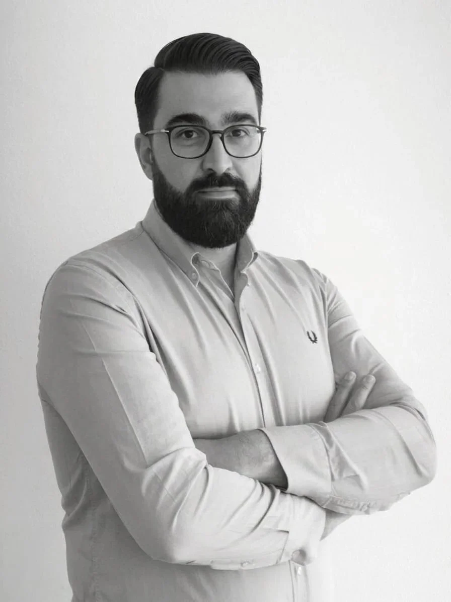 Uomo con barba, occhiali e camicia a maniche lunghe, con le braccia incrociate, in un ambiente bianco.