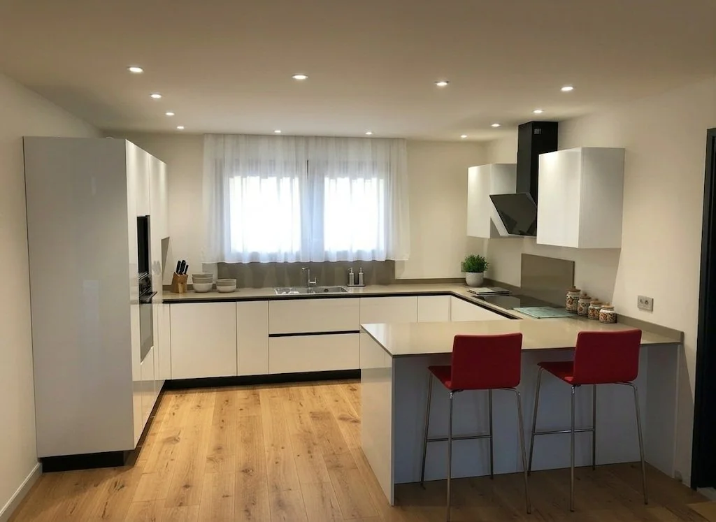 Cucina moderna con ante bianche, bancone con due sedie rosse, lampade sul soffitto, piante e stoviglie sulla finestra.