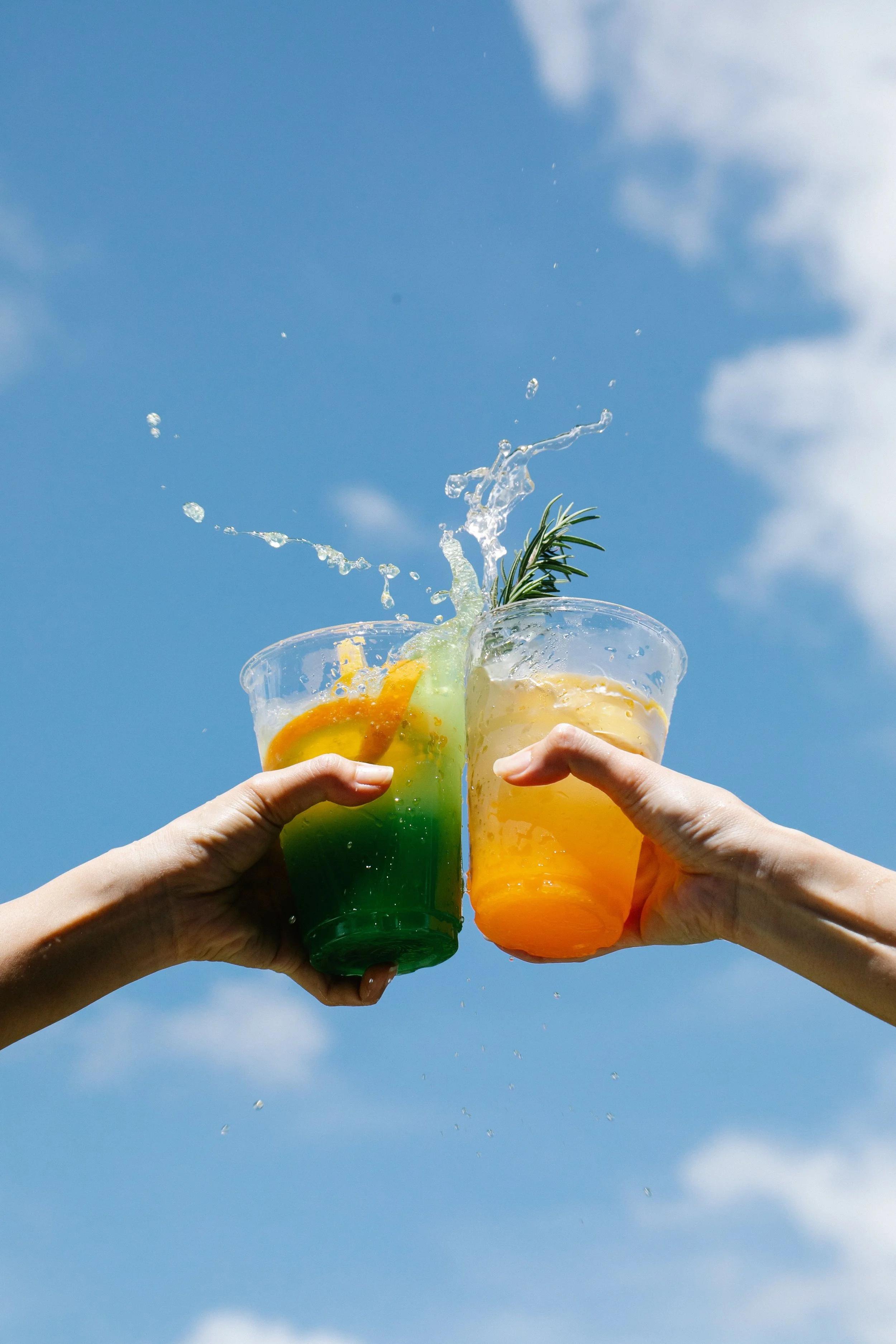 Two hands holding colorful drinks with fruit garnishes under a blue sky with clouds, with water splashing out of the drinks.