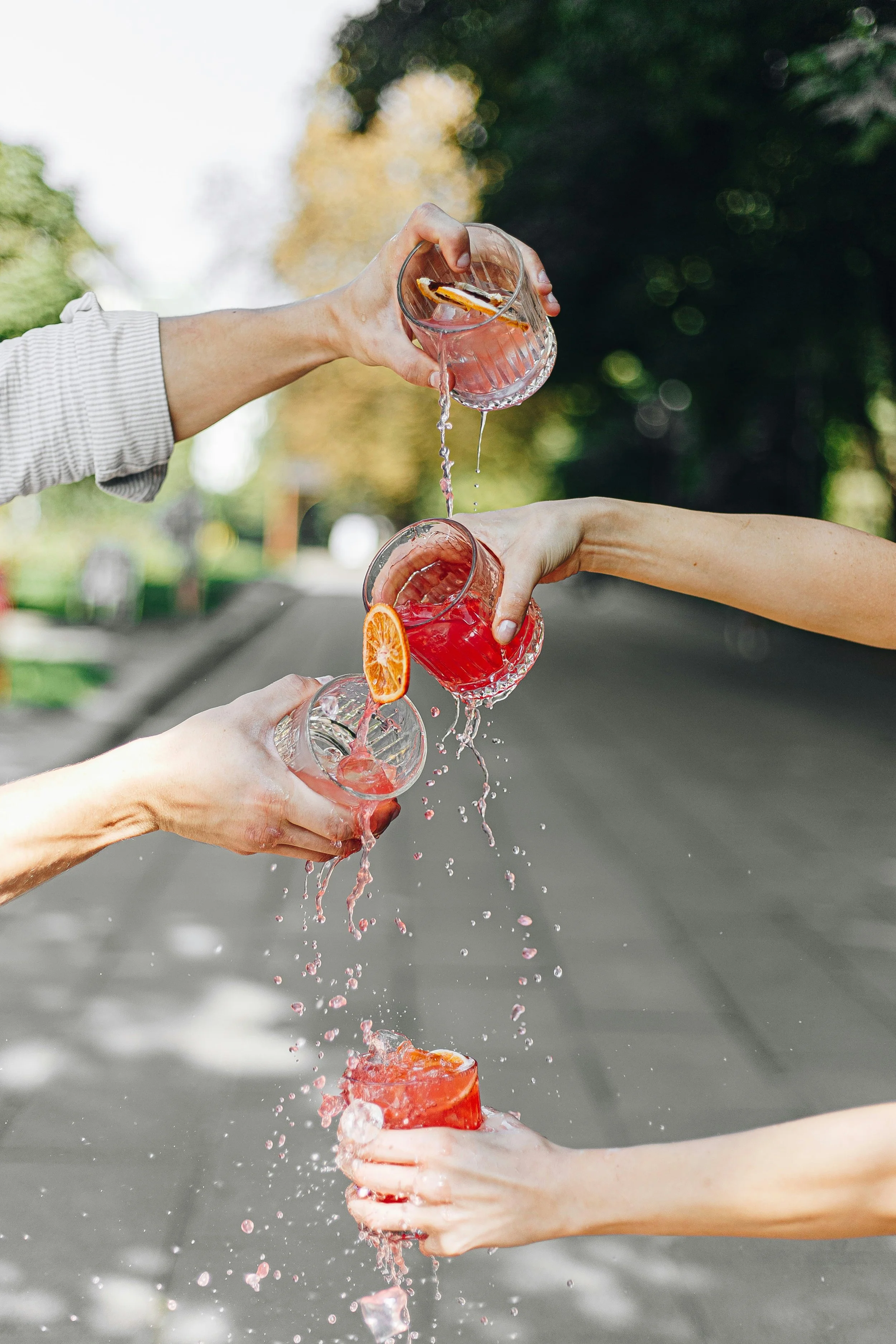 Multiplehands are holding glasses with drinks, some garnished with orange slices, as they pour liquids into each other's glasses outdoors with blurred trees and a road in the background.