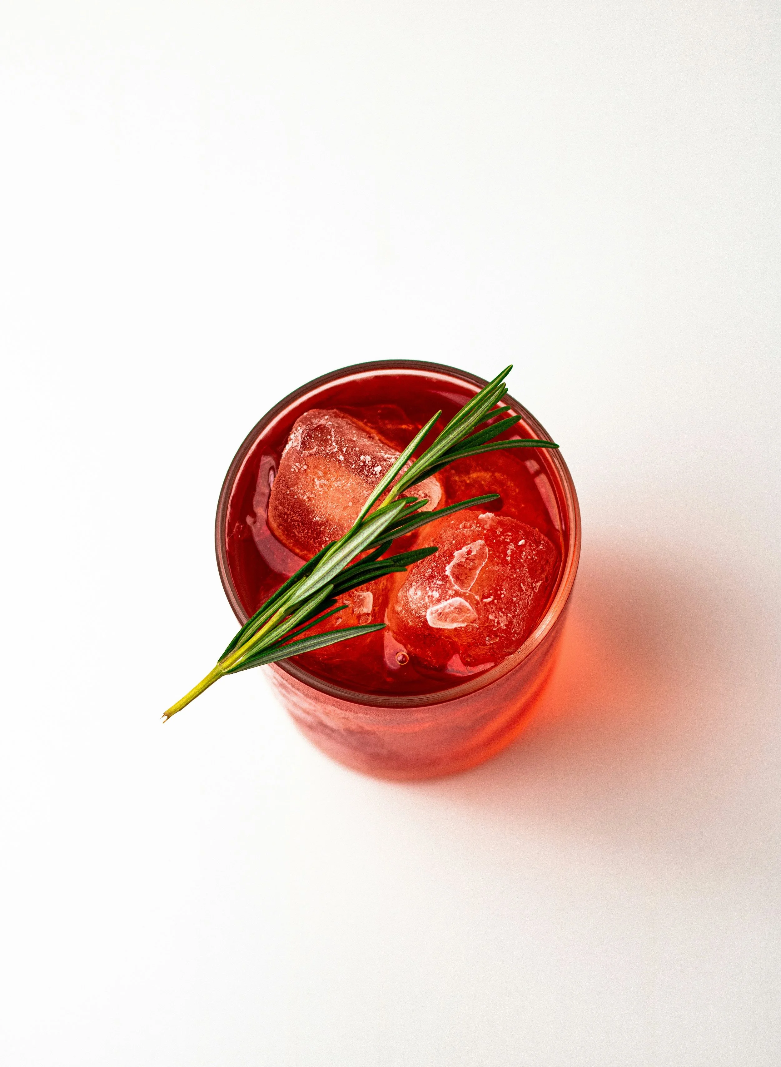 Red glass of a drink garnished with a sprig of green herbs and ice cubes, viewed from above on a white surface.