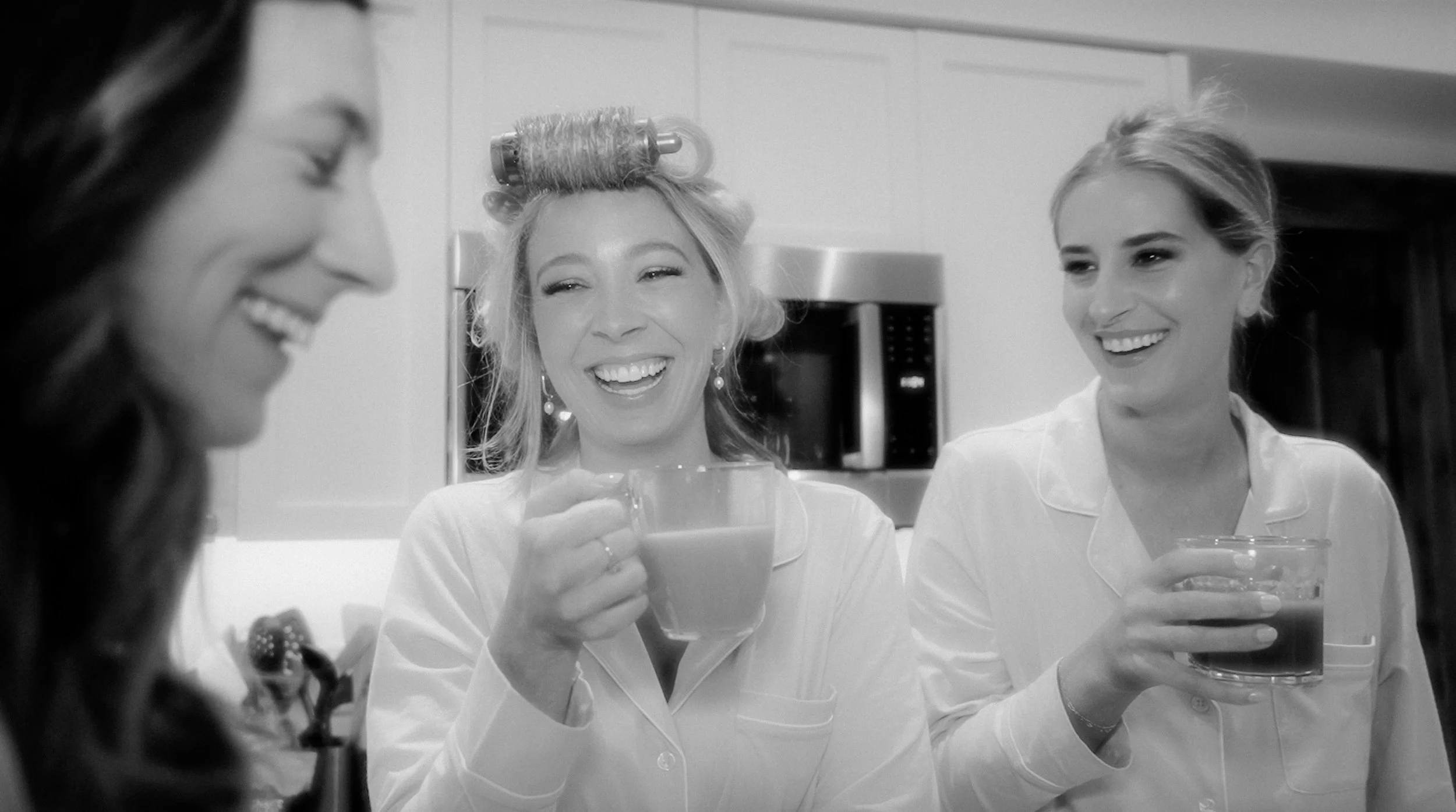 Three women enjoying drinks and laughing, one woman with a hair roller, in a kitchen setting.