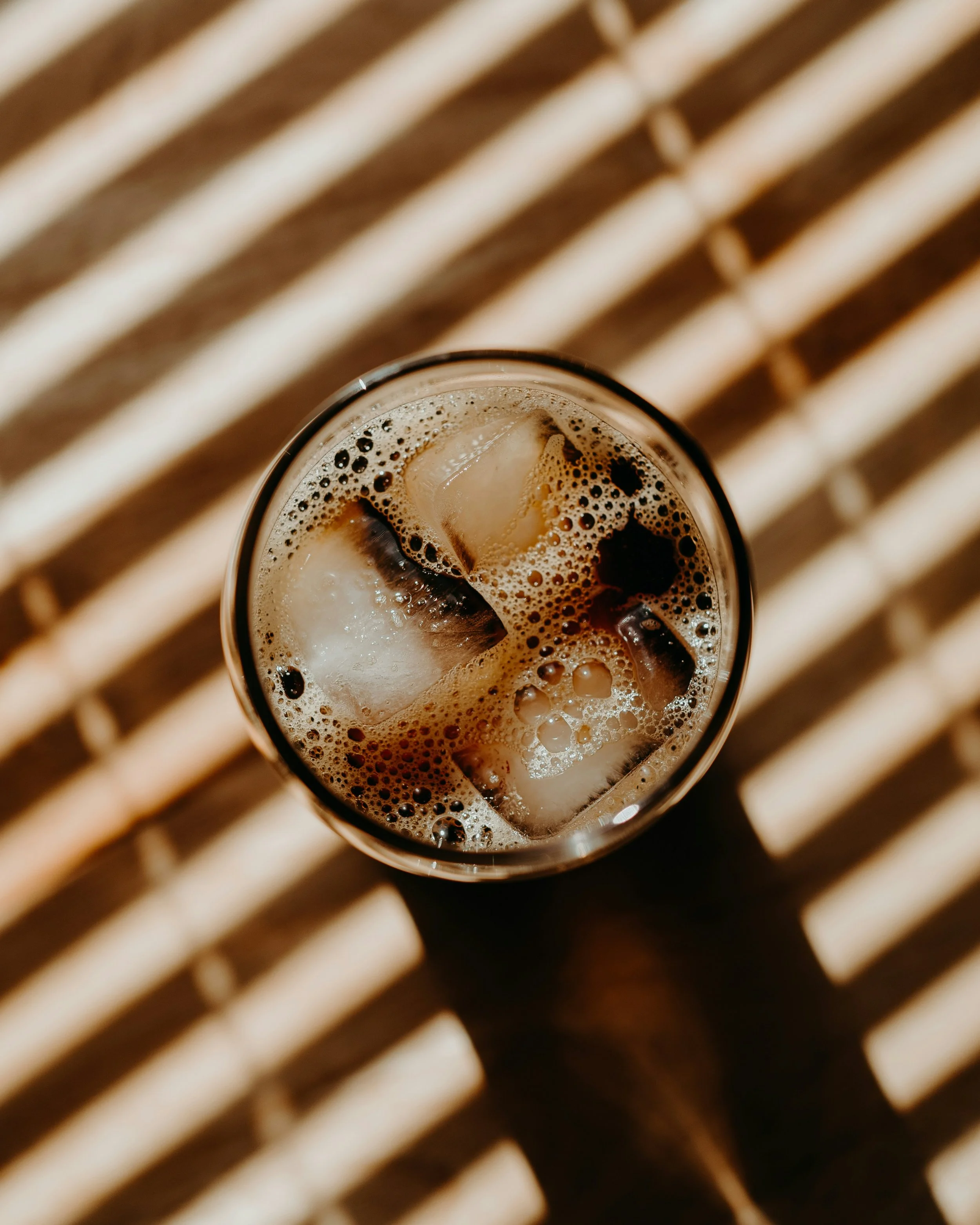 A glass of iced coffee with ice cubes, seen from above, on a striped surface with alternating light and dark lines.
