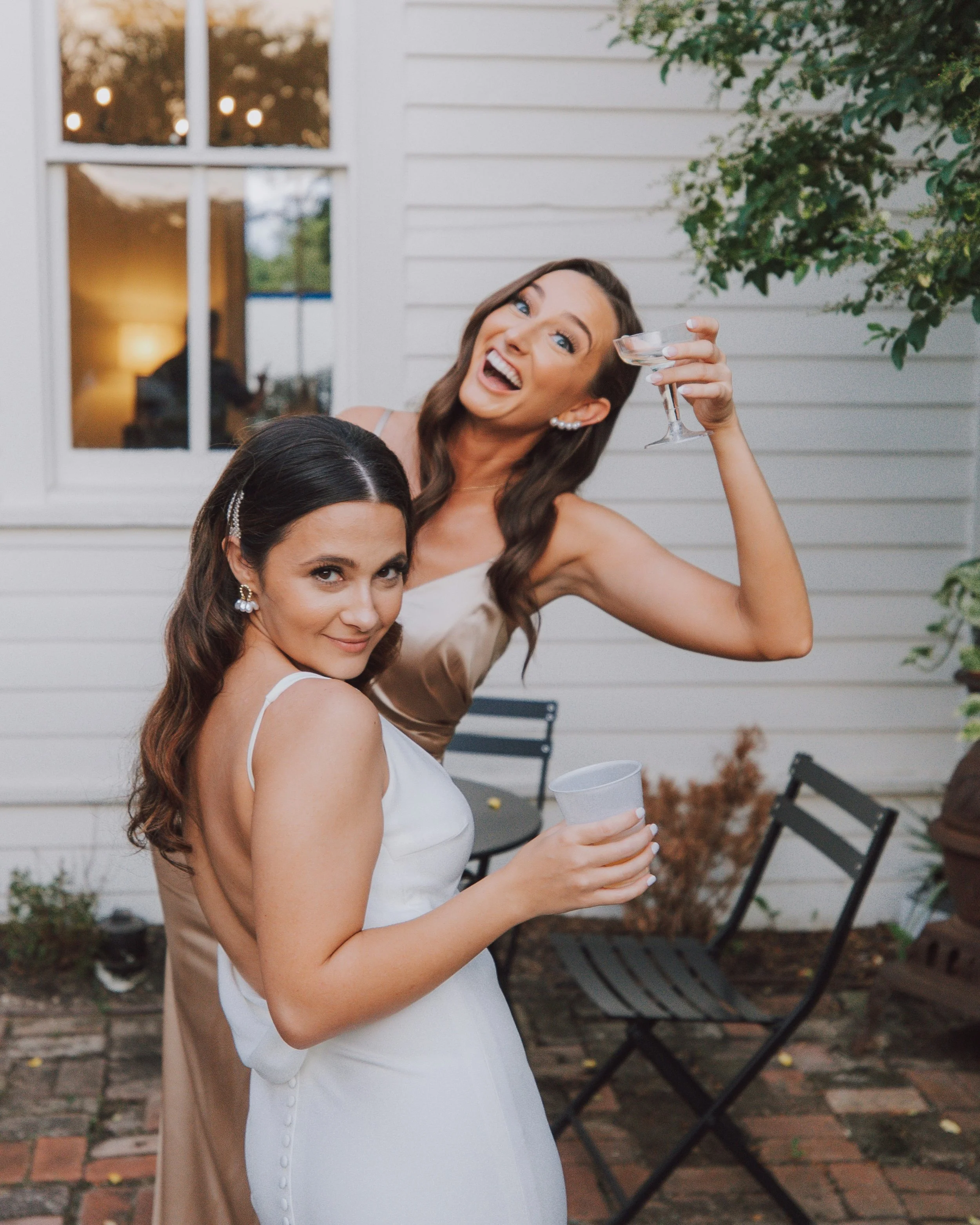 Two women celebrating outdoors. The woman in the foreground has long dark hair, a white dress, and pearl earrings. The woman behind her has medium-length brown hair, a beige dress, and is holding a glass of wine, smiling. They stand on a brick patio with outdoor furniture and a white house with a window and greenery in the background.