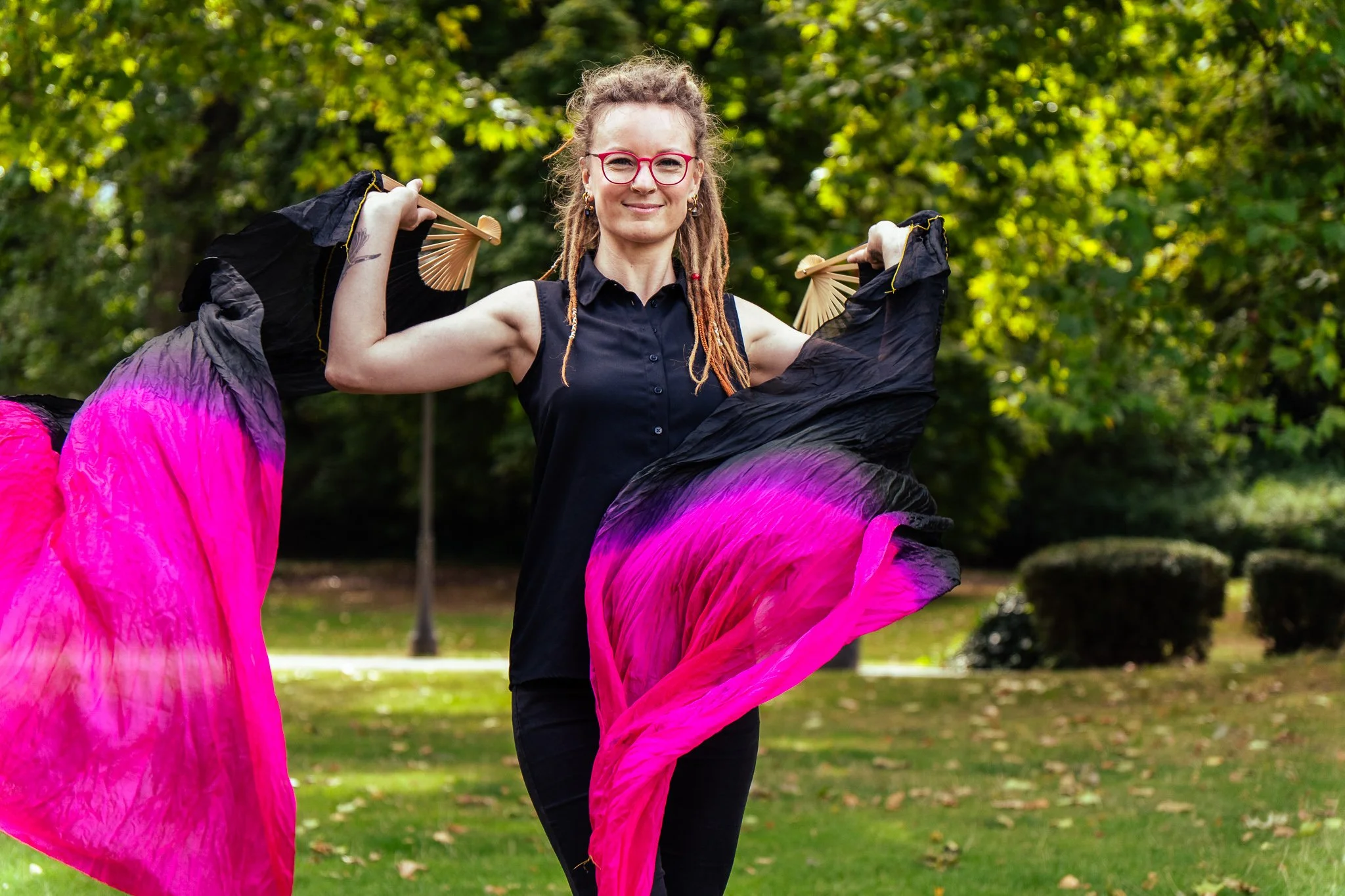 Eine Frau mit dreadlocks, roter Brille und schwarzen Kleid tanzt mit bunten Stoffflaggen im Park.