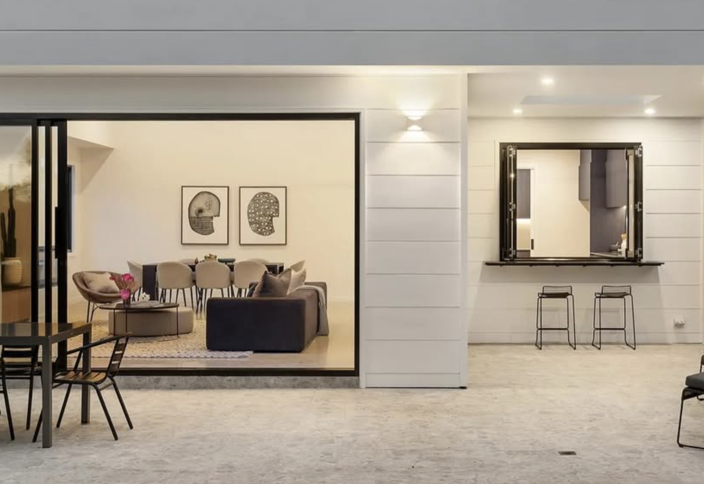 A modern, open living space with a secondary outdoor bar area, white paneled walls, and minimalistic furniture including bar stools, a dining table, and artwork.