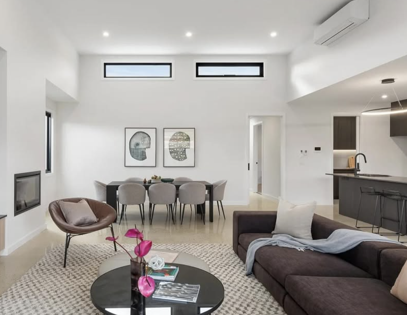 Modern, open-concept living room and dining area with white walls, high ceiling, and minimal decor. Features a brown sofa with a white pillow, a round black coffee table with pink flowers, a modern armchair, a dining table with eight chairs, and framed abstract art on the wall. Kitchen with black cabinets and a breakfast bar is visible in the background.