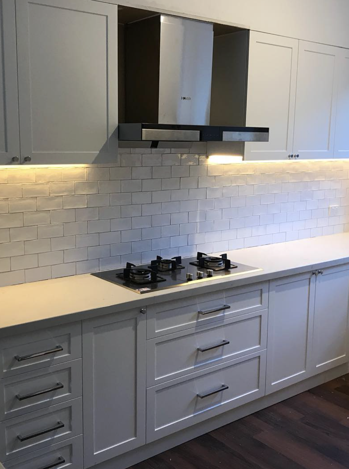 White kitchen cabinet with drawers and a gas stovetop above a white countertop, beige subway tile backsplash, wall-mounted stainless steel range hood, under-cabinet lighting, dark wooden floor.