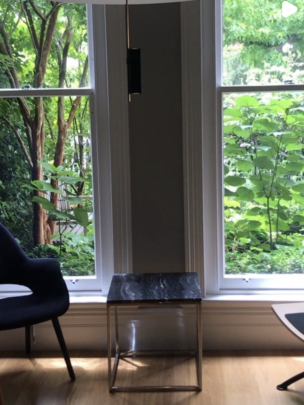 A room with large windows showing green foliage outside, a black armchair on the left, a small black marble-top table in the center, and part of a desk or table on the right.