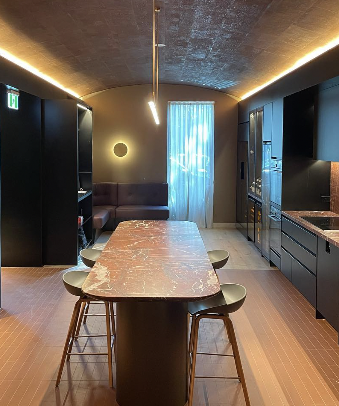 Modern kitchen with wooden dining table, round chairs, black cabinetry, blue curtains, and warm ambient lighting.