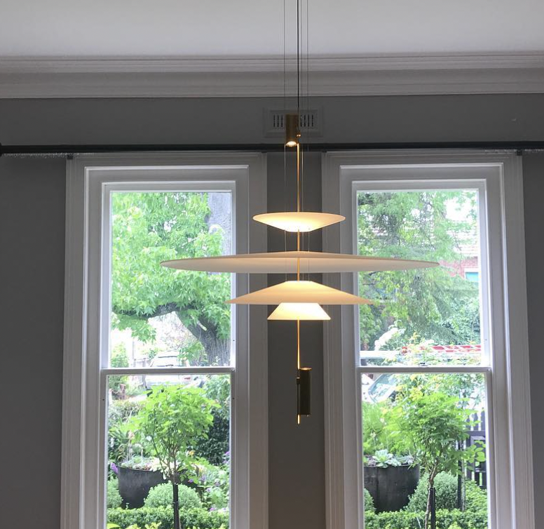 Modern hanging light fixture with three circular, disc-shaped shades hanging in front of two large windows with greenery outside.