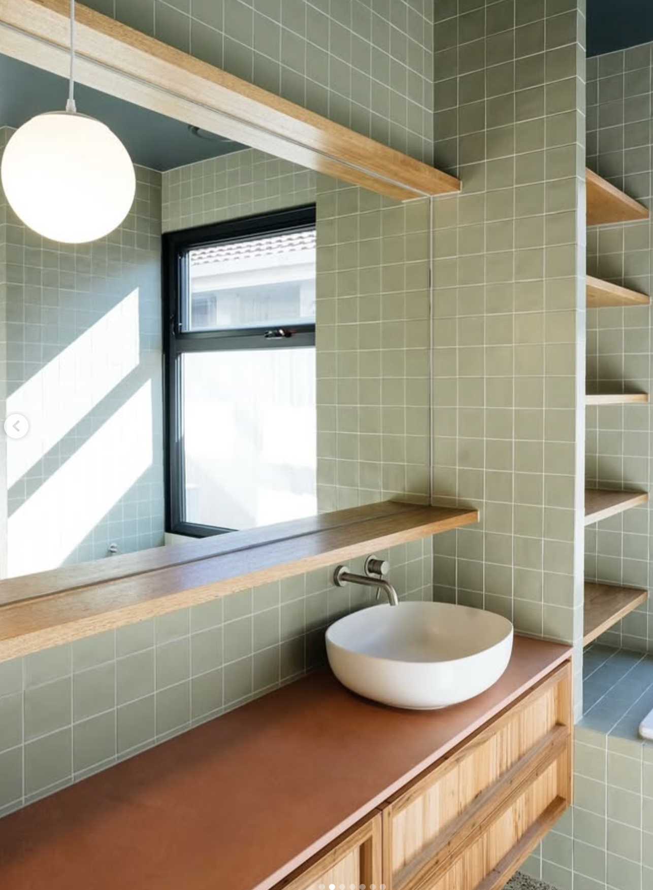 A modern bathroom with green tiled walls, a large mirror, a round white vessel sink, wooden shelves, and a window letting in natural light.