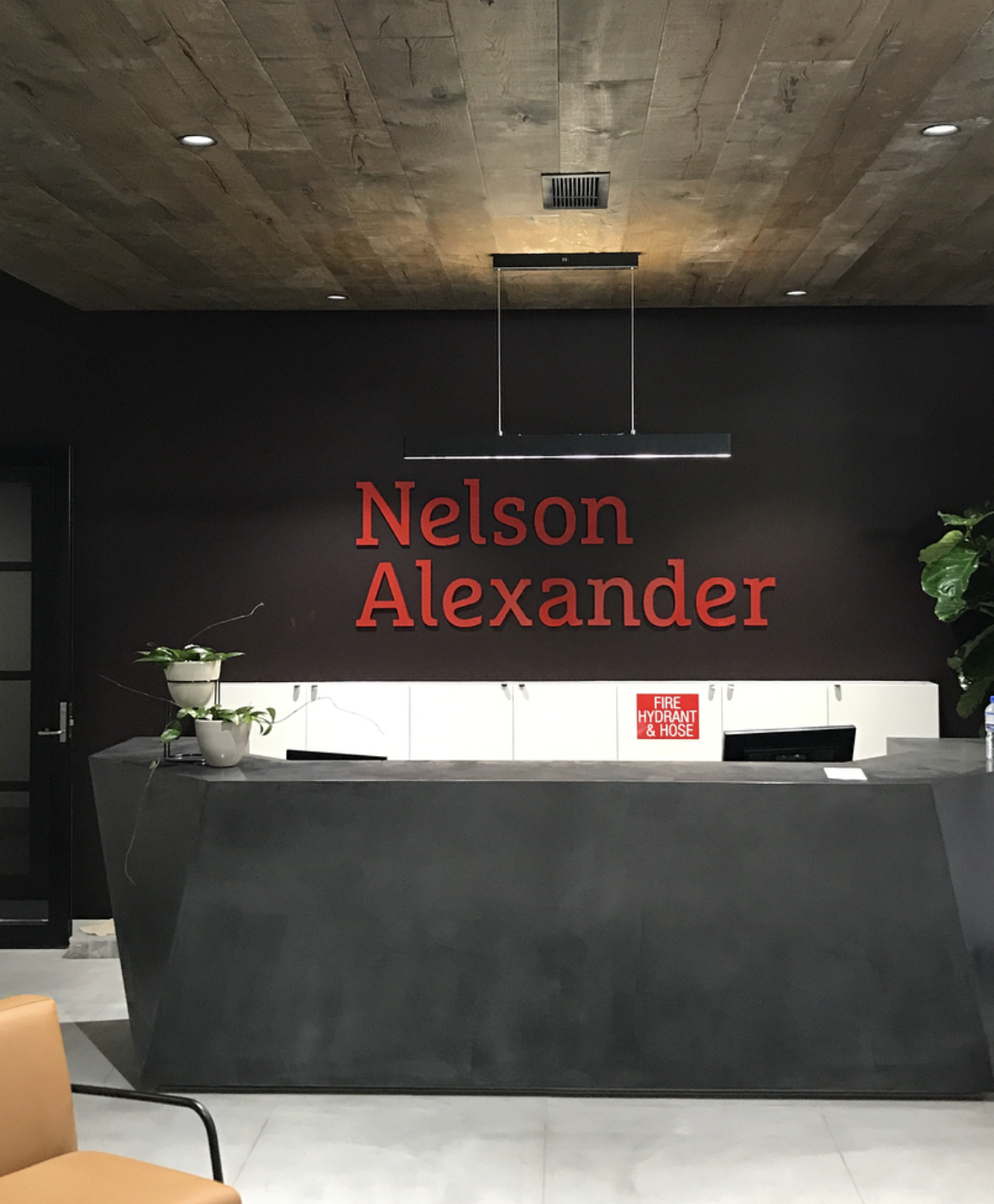 Reception desk with plants and a name sign that reads 'Nelson Alexander' on a dark wall, with a fire hydrant and hose sign below.