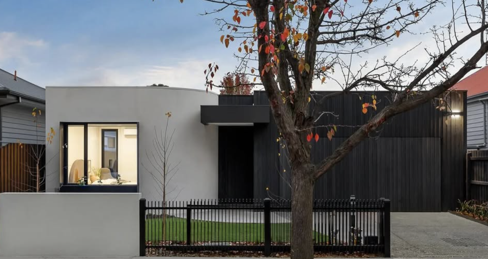 Modern house exterior with a large front window, a small tree in front, and a big tree with fall leaves in the yard, all enclosed by a black fence.