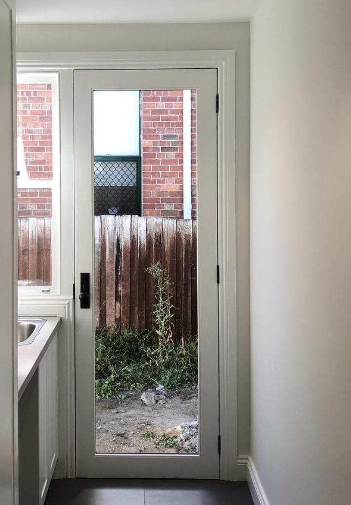 View through a door with a glass panel showing a small backyard with a wooden fence, some weeds, and red brick exterior walls.