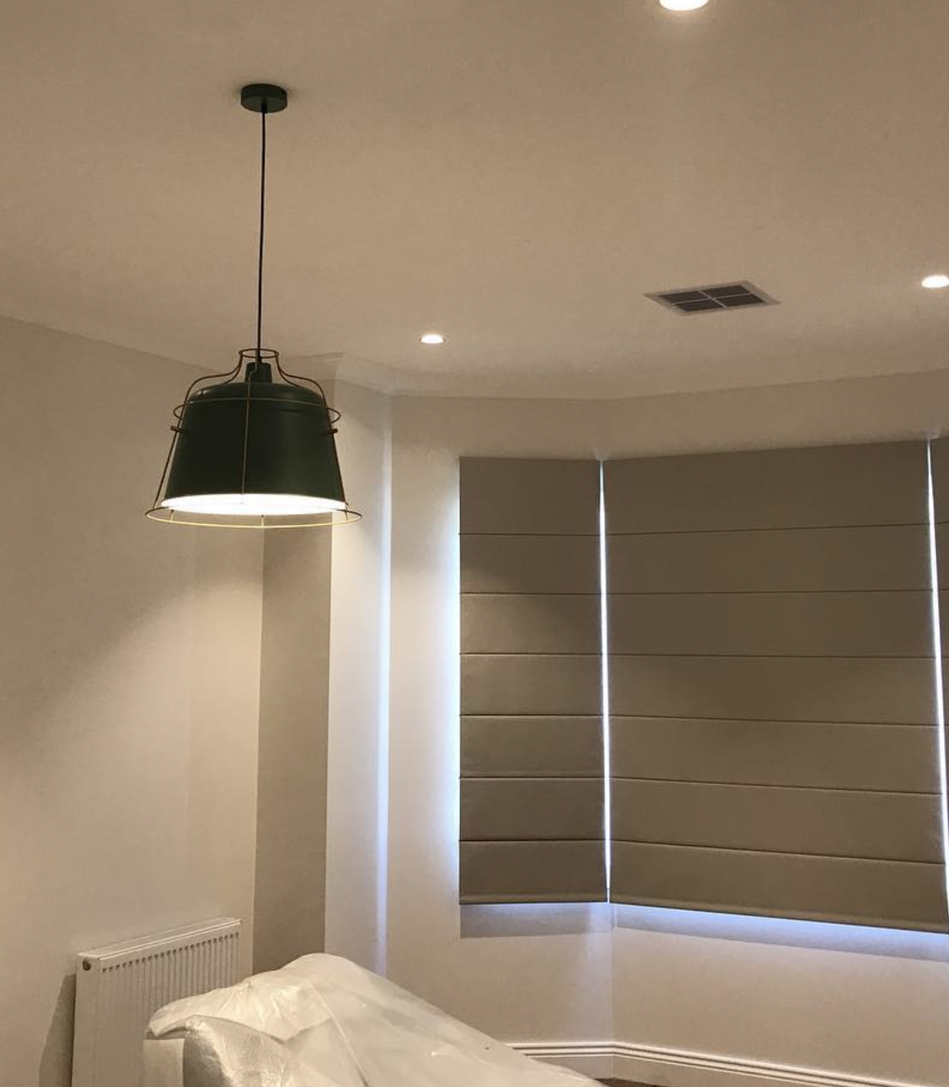 Interior view of a bedroom showing a modern black pendant light hanging from the ceiling, a window with beige blinds, a white radiator, and part of a bed with white bedding in the corner.