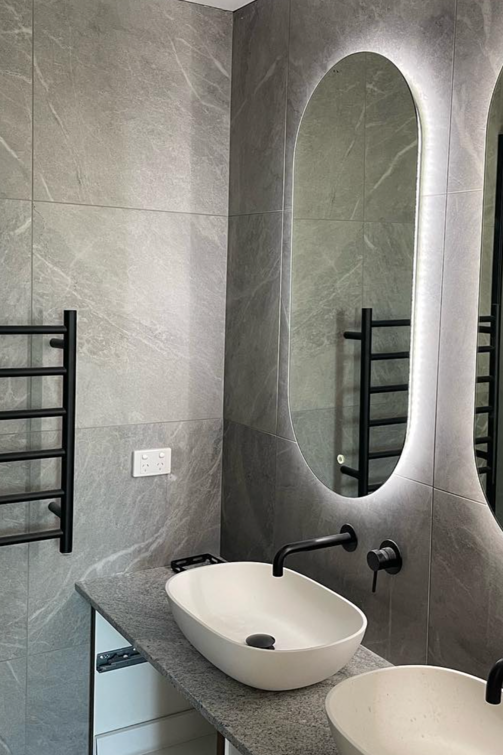 Modern bathroom with gray tiled walls, a wall-mounted black faucet above a white vessel sink on a gray countertop, mirrored wall with LED lighting, and a black heated towel rail on the wall.