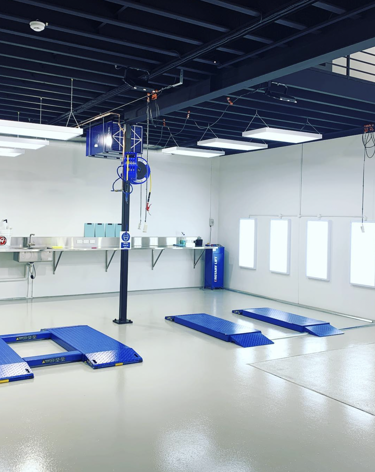 A clean automotive workshop with blue vehicle lifts on a white floor, bright white wall with windows, and a workbench with tools and equipment.