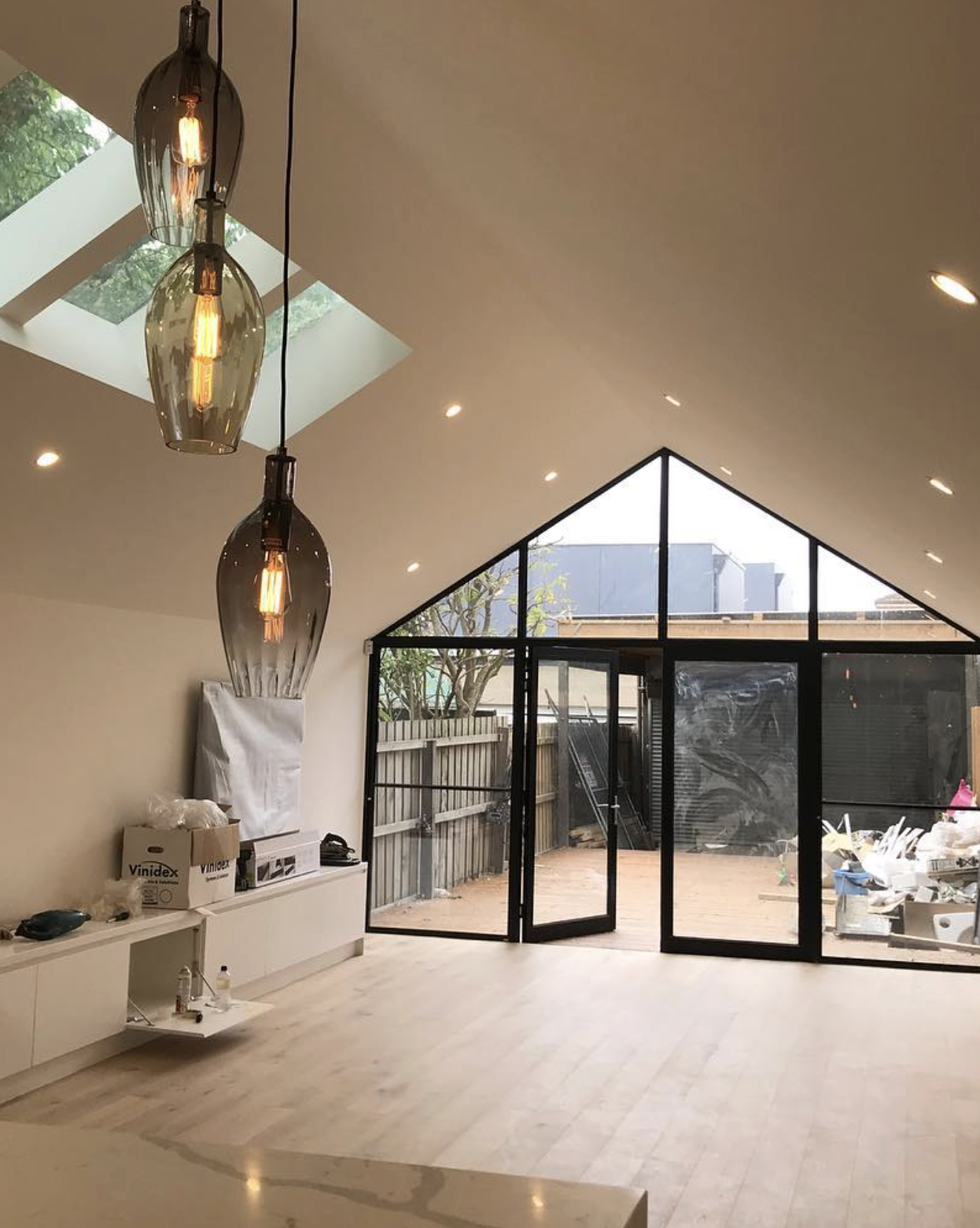 Modern living room with pendant lights, skylight, and large windows opening to an outdoor patio with construction materials.