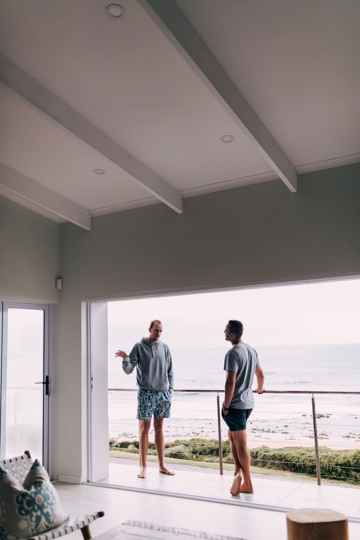Two men talking on a balcony overlooking the ocean, one in a hoodie and shorts, the other in a t-shirt and shorts, inside a room with a sliding glass door and a ceiling with beams.