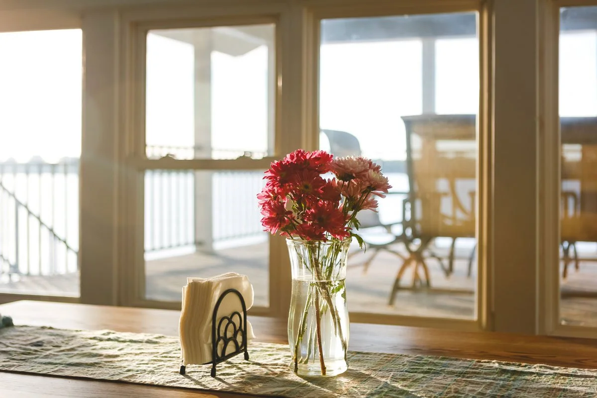 A glass vase with pink and white flowers sits on a wooden table near a window, with a balcony and outdoor furniture visible in the background.