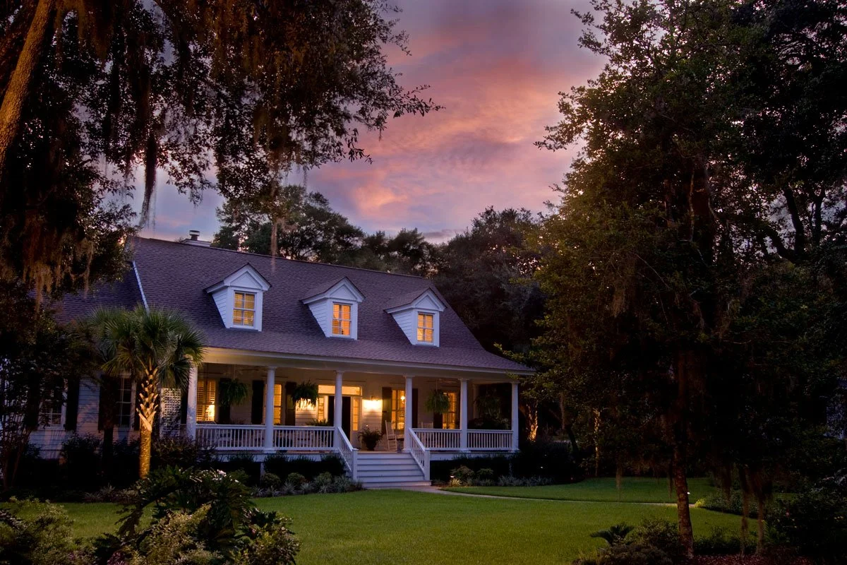 A two-story house with a porch and white railings, surrounded by trees and greenery, at sunset with the sky painted in pink and purple hues.