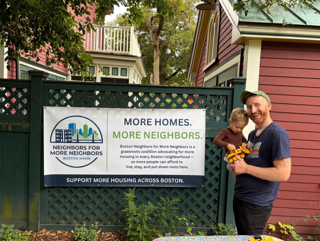 A smiling man holding a young girl with a toy truck, standing next to a sign that promotes affordable housing and community support in Boston. There are houses and trees in the background.