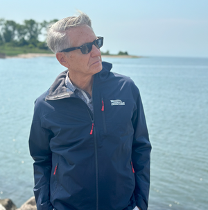 Older man with gray hair wearing sunglasses and a navy North Face jacket standing by a body of water with a distant shoreline.